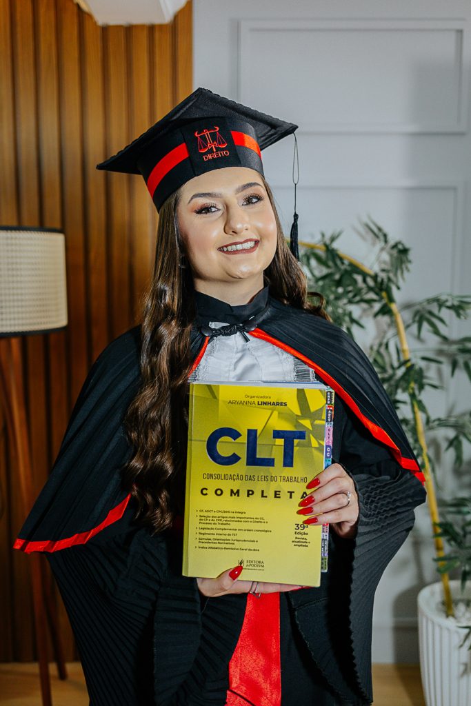 Formanda de Direito em Curitiba posando com beca preta, faixa vermelha e livro CLT em ensaio de estúdio.