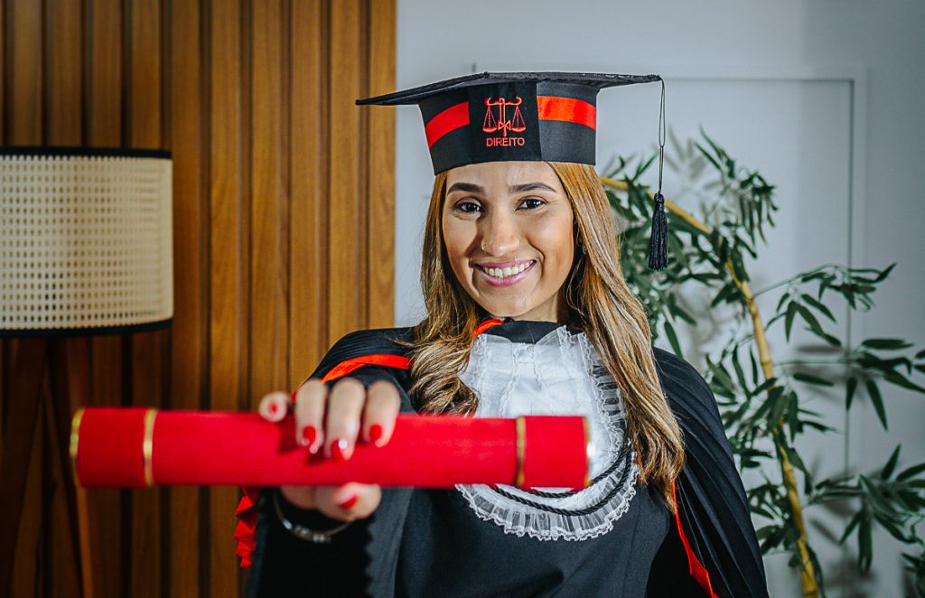 Formanda de Direito posando com beca e faixa vermelha em fundo neutro de estúdio.